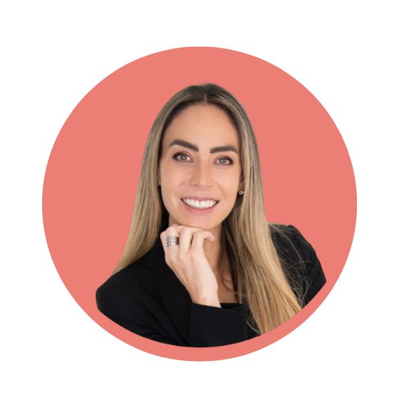Smiling woman with long hair wearing a black blazer, resting her chin on her hand against a coral circular background.