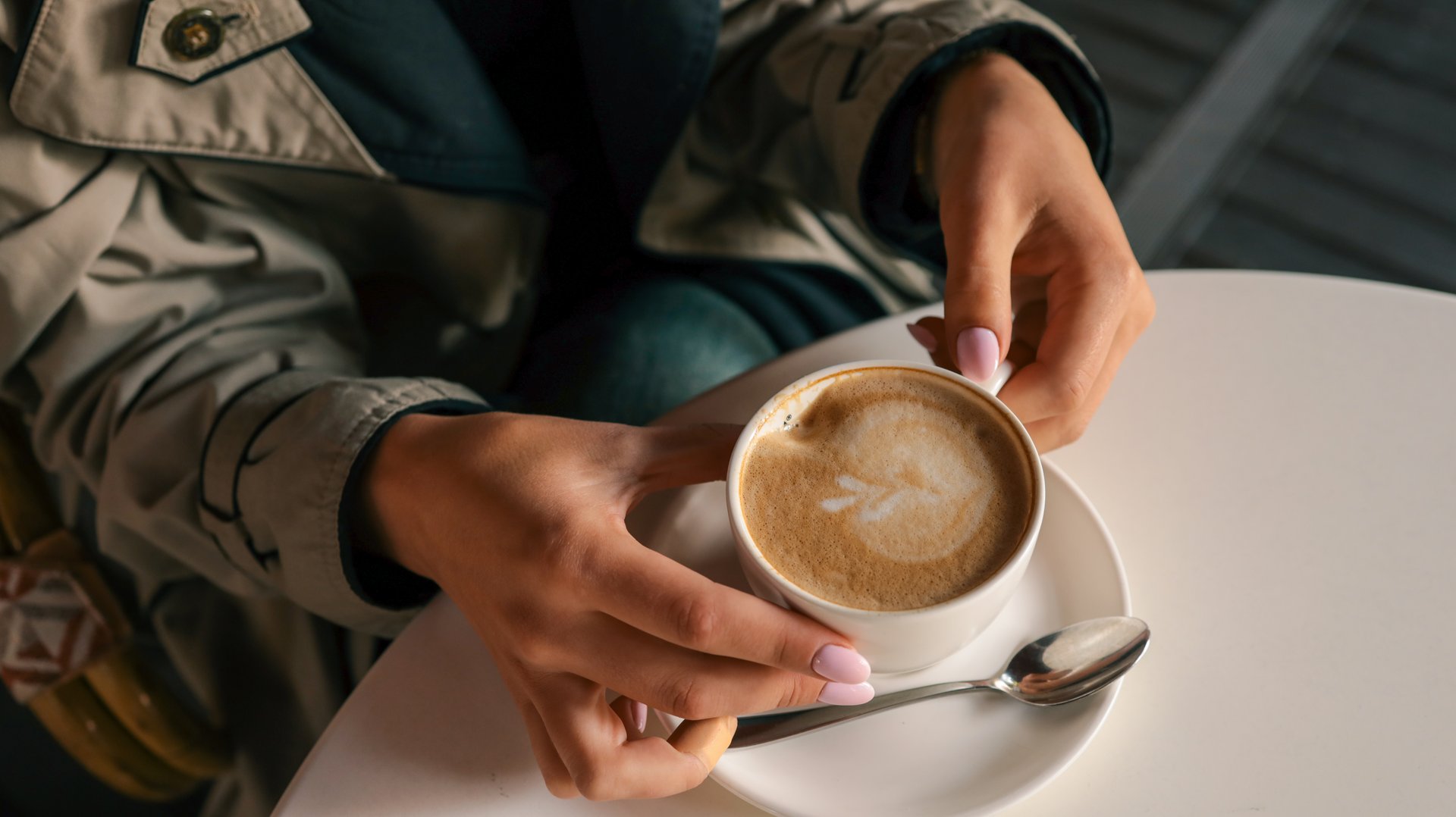 Close-up of womanâs hands in trench coat gently holding a cup of cappuccino with latte art, seated at a white table in a cozy outdoor cafÃ© setting.