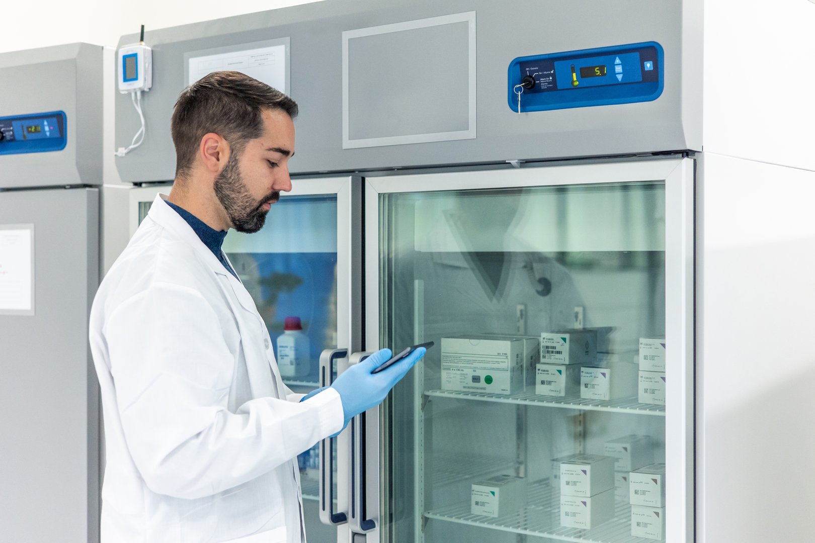 Scientist wearing a lab coat and gloves is using a digital tablet to check inventory or record data related to medical samples stored in a specialized laboratory refrigerator