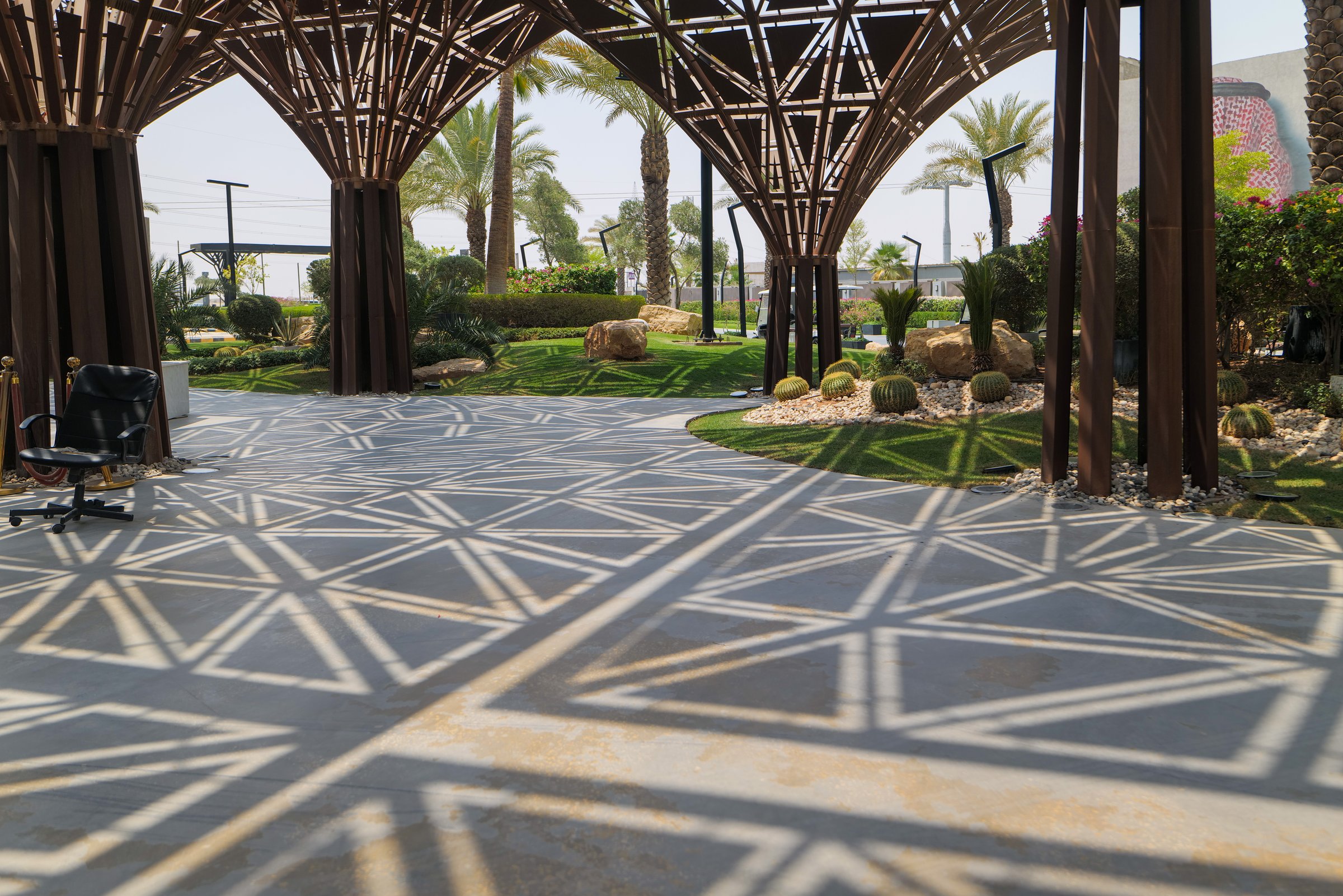 Architectural Pergola and Desert Garden in Boulevard, Riyadh. An artistic photo showing a modern pergola with an intricate geometric design, casting striking triangular shadows on the paved ground. The structure frames a beautifully landscaped garden featuring native desert plants like cacti and palm trees, with large rocks and green grass creating a serene and contemporary outdoor space. High quality photo
