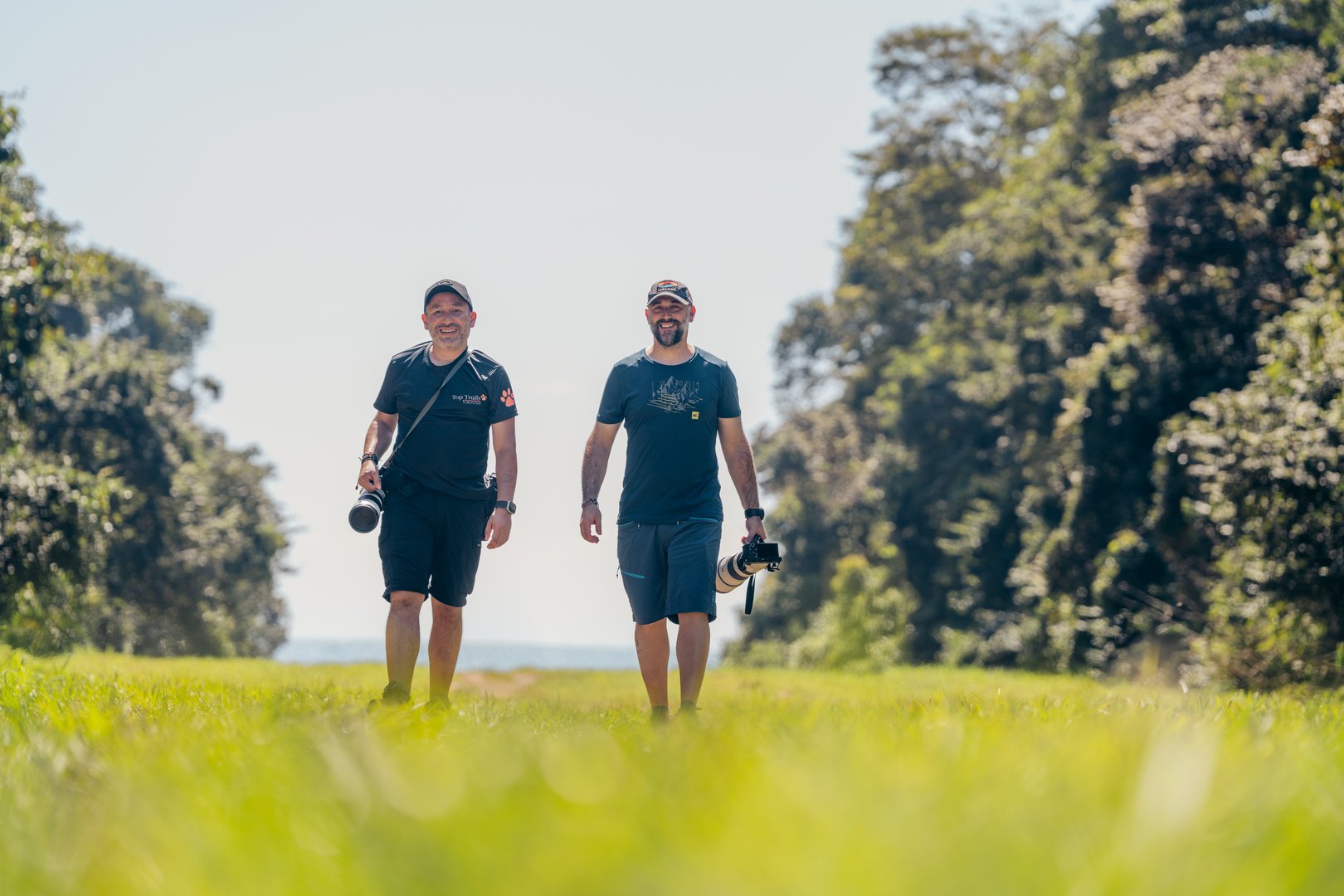 Two men walk through a grassy field with cameras, flanked by trees in the background, under a clear sky.