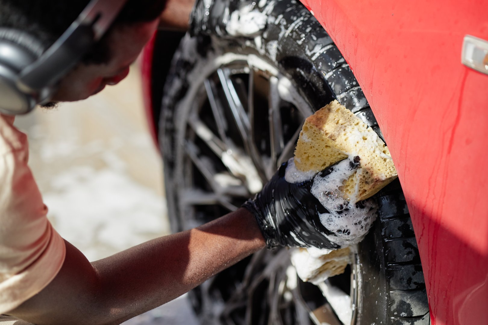 Black man wearing headphones cleaning car wheel with soapy sponge, focusing on scrubbing tire surface, close up of gloved hand working on red vehicle during car wash