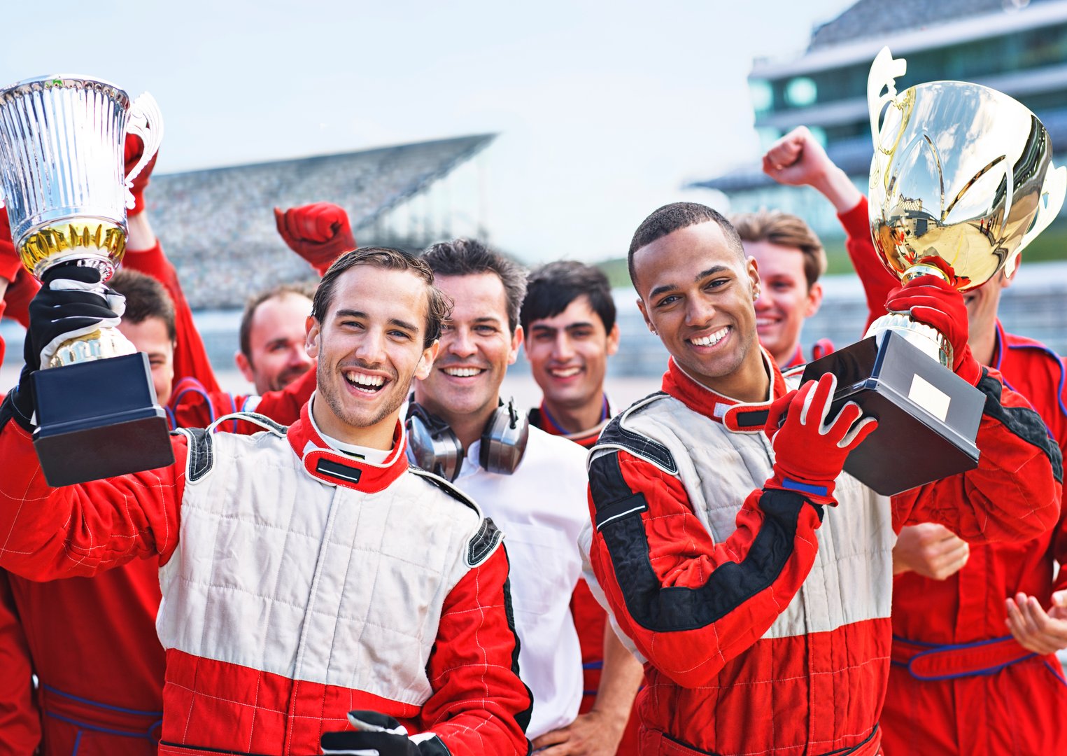 Two race car drivers in red suits celebrating with trophies, surrounded by a cheerful team at a race track.