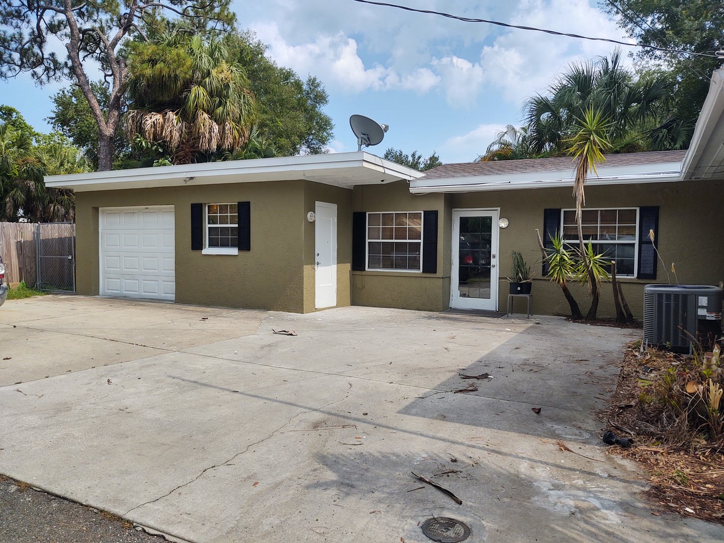 Single-story house with green exterior, white garage door, and satellite dish on the roof. Surrounded by palm trees.