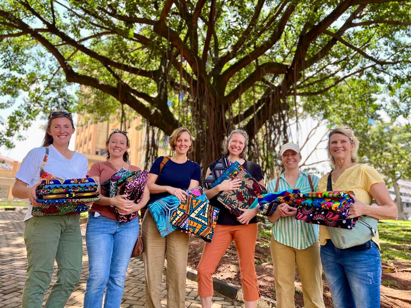 Six smiling women holding colorful fabrics, standing under a large tree in a sunny outdoor setting.