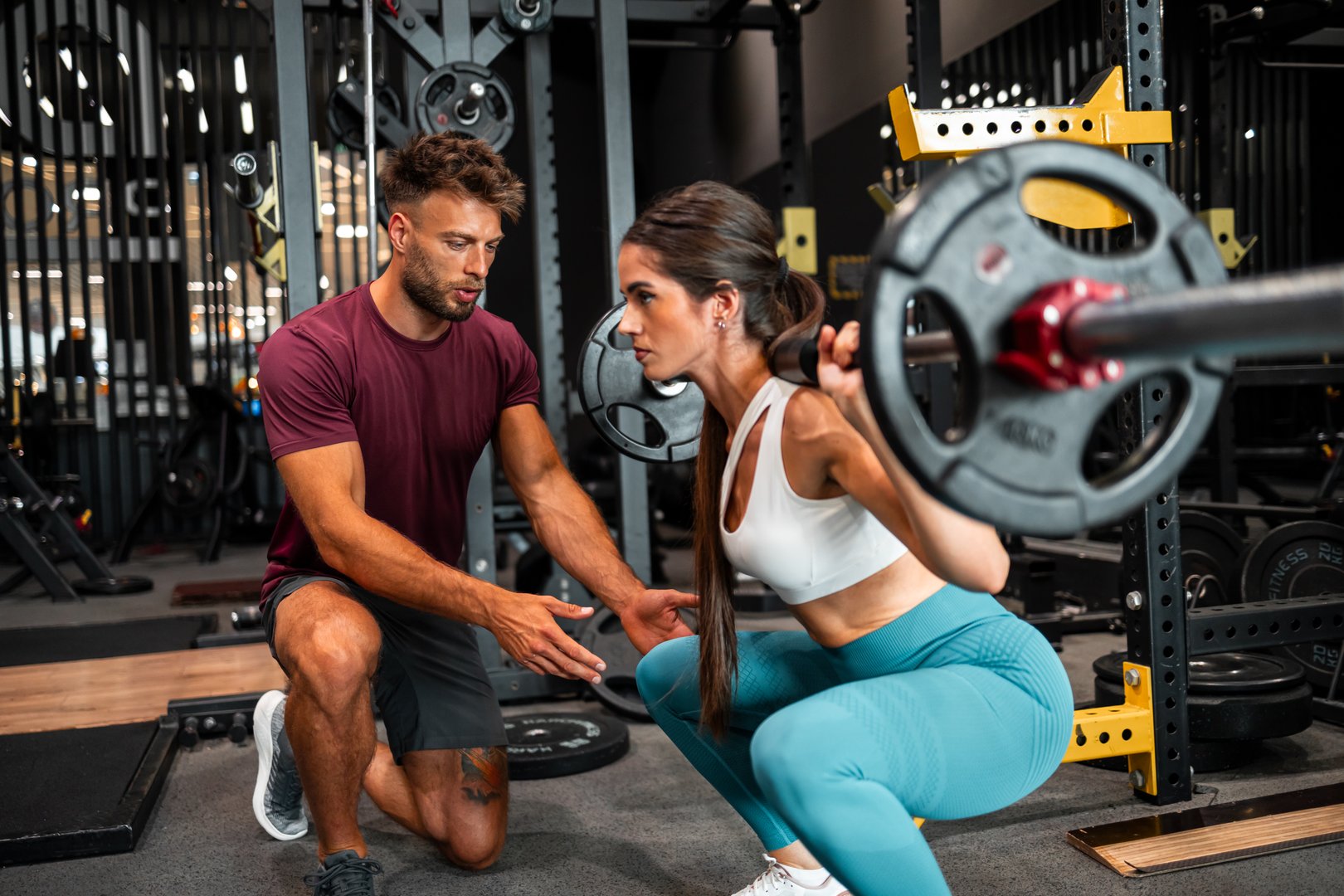 A personal trainer instructs a client performing squats with weights at a professional gym setup. The interactions demonstrate dedication, fitness training, teamwork, and working towards health and body strength goals.