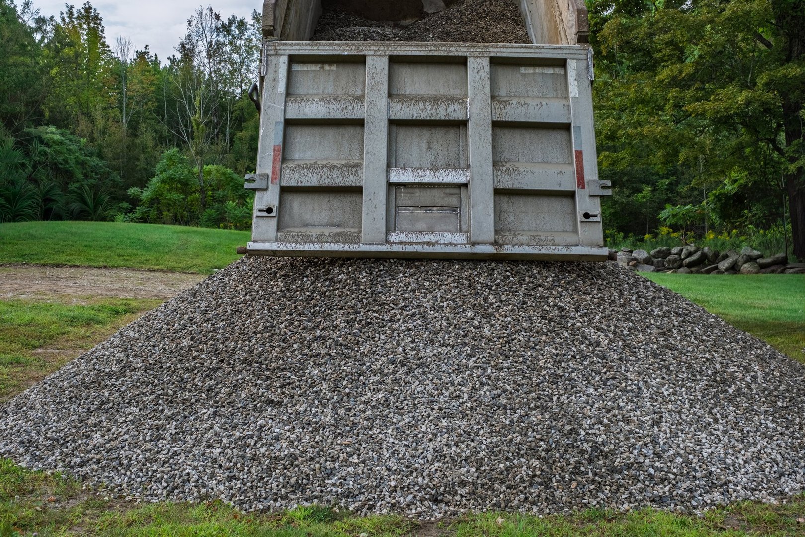 A dump truck unloads a large pile of crushed stone onto a grassy yard