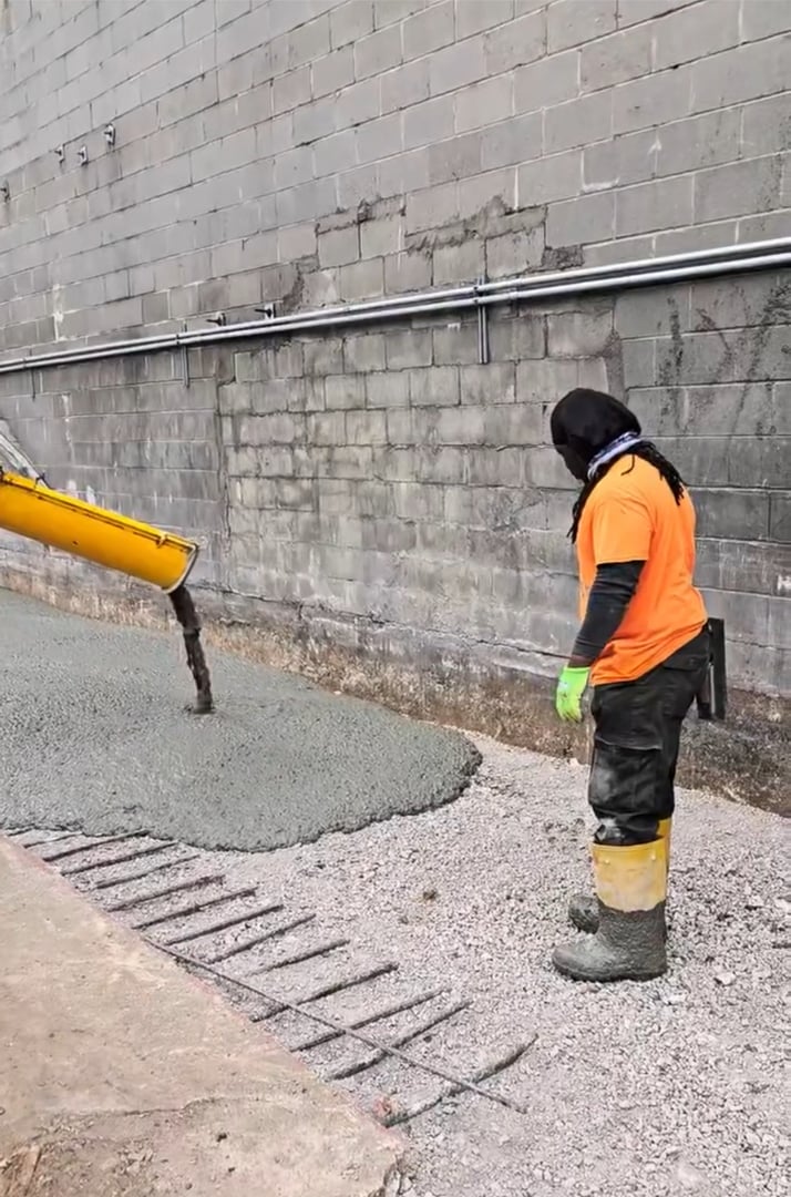 Worker with orange shirt and yellow boots supervises pouring concrete on gravel near a brick wall.