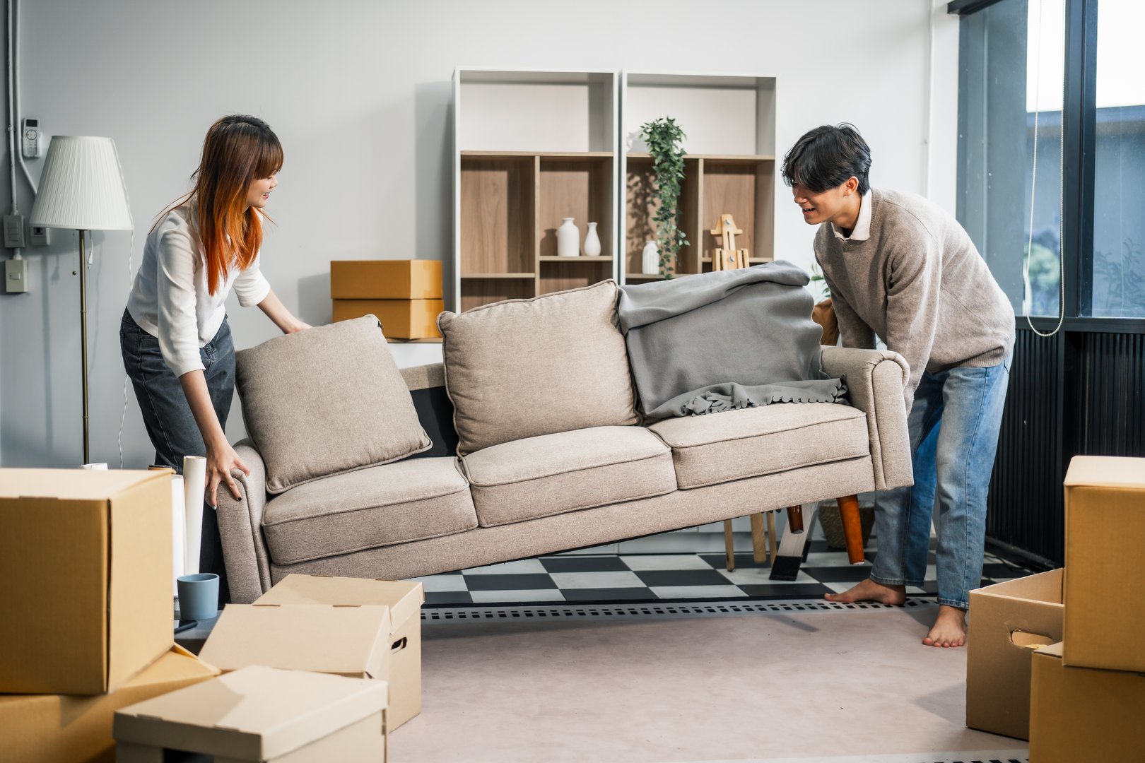 A young couple happily moves furniture together in their new home. They work as a team, lifting and carrying boxes and belongings, creating a happy, familial atmosphere.