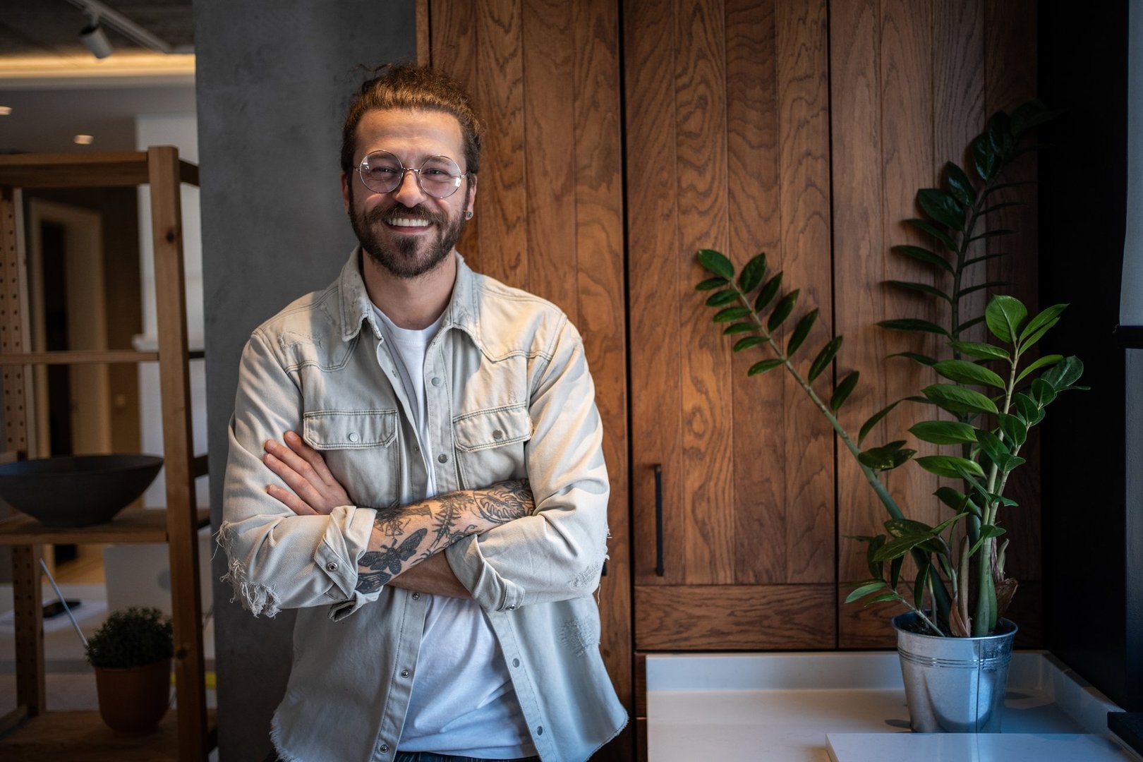 Portrait of a cheerful young man with crossed arms and tattoos, standing in a modern kitchen