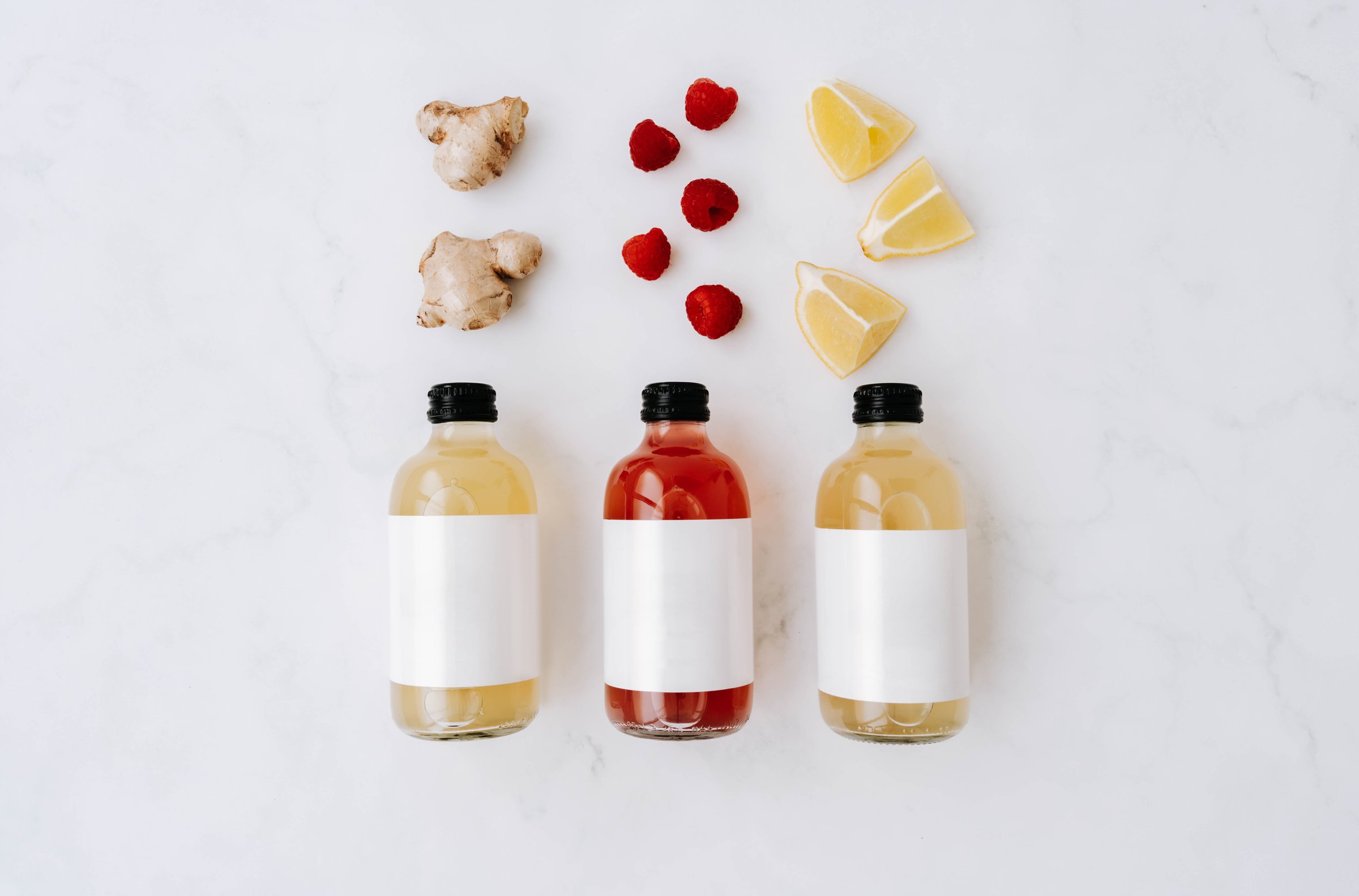 Three small transparent glass bottles with black caps, filled with colorful kombucha of yellow, red, and green and ingredients on a white surface. Each bottle has a plain white label. Top view. Copy space