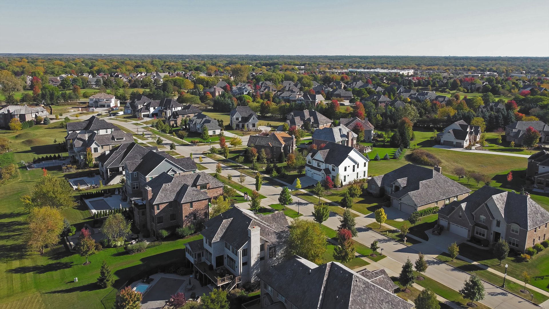 Aerial view of suburban homes in Chicago, Illinois. An expansive aerial view of a suburban area in Chicago, Illinois, showcasing large residential homes with landscaped lawns, tree-lined streets, and autumn foliage under a clear sky.
