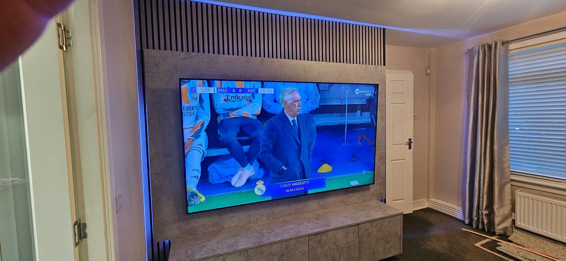 A TV displaying a soccer match with a coach standing near the sidelines. Living room setting with curtains and a door.