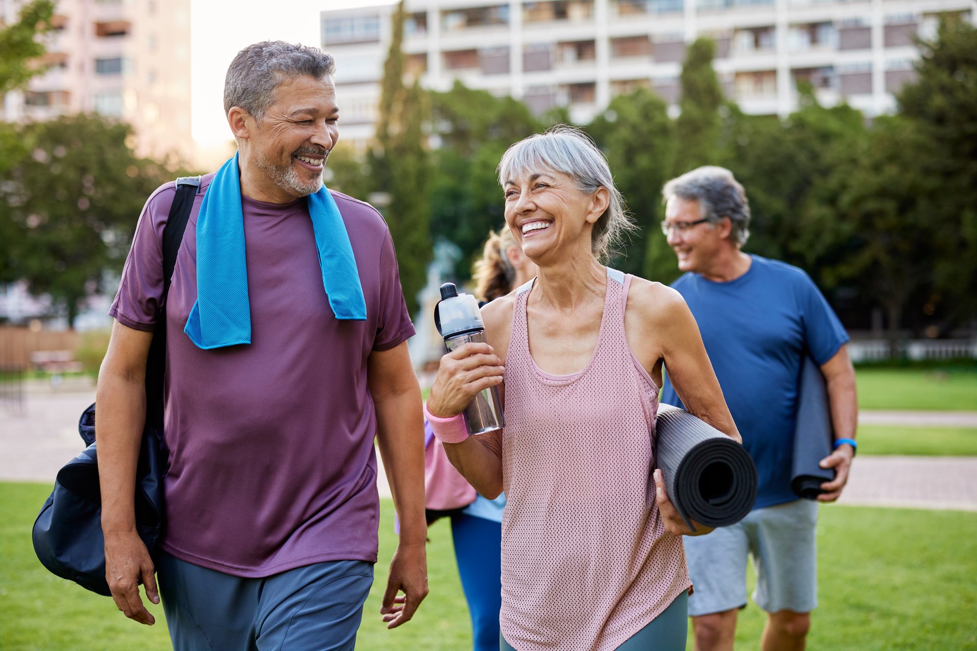 Happy elderly couple walking after yoga in a city park. Group of senior men and women smiling and talking with yoga mats and water bottles after sport exercise. Group of older adults enjoying fitness and friendship outdoors at park.