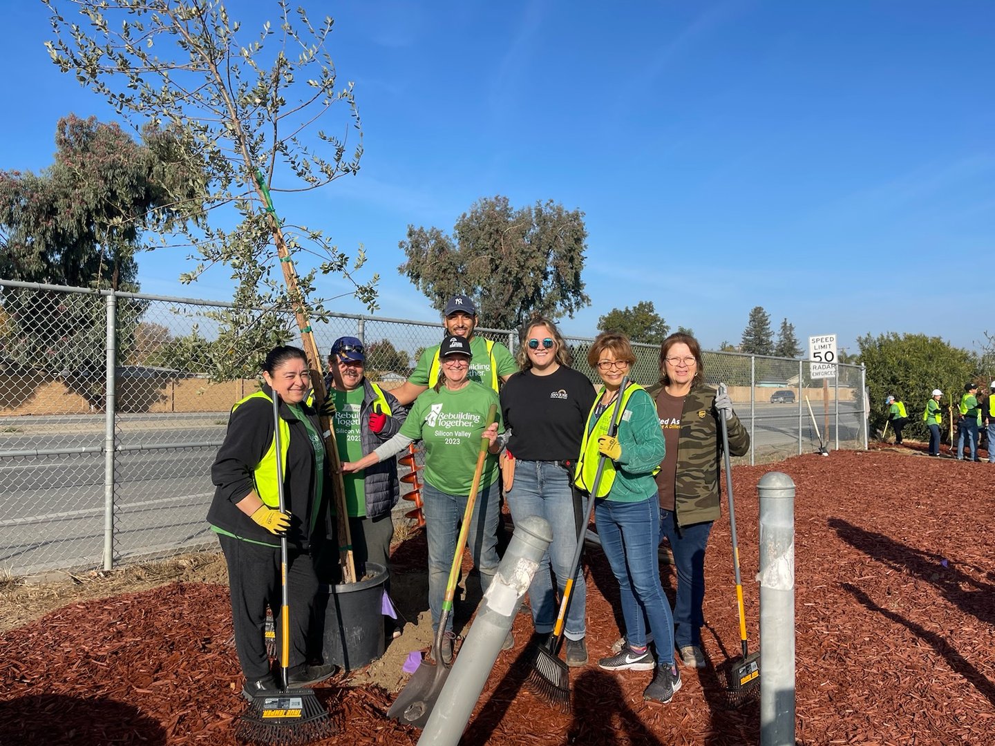 Group of people planting a tree beside a fence, wearing green shirts and vests, holding shovels and smiling at the camera.