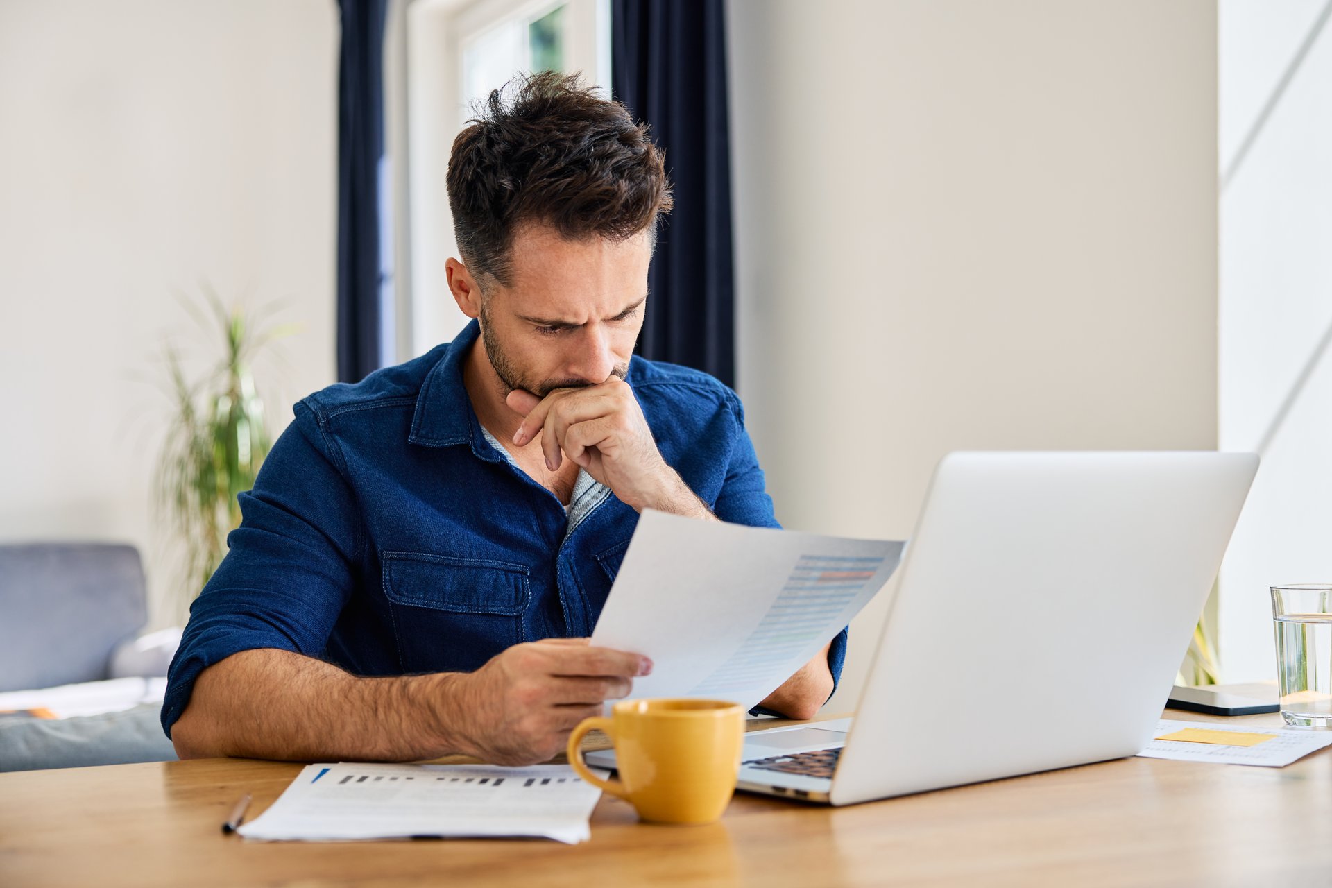 Sad man looking at financial documents at home sitting at a table with laptop in living room