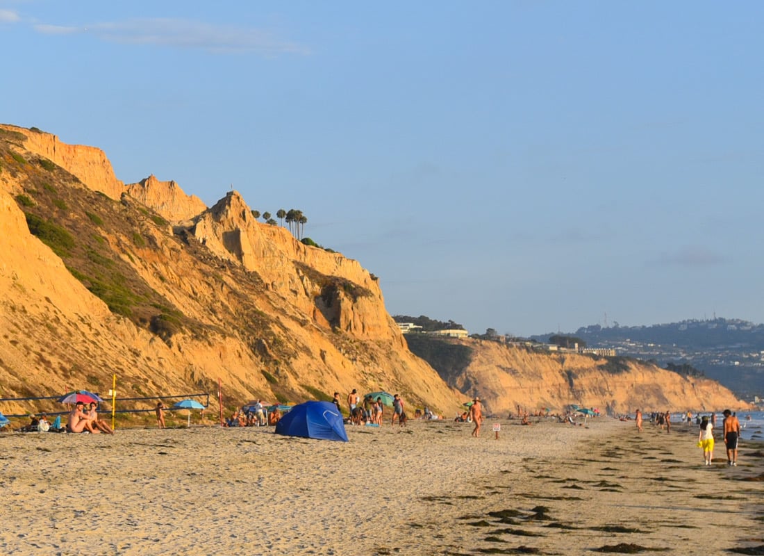 Clifftop view of a beach with people, a blue tent, and rugged cliffs in the background under a clear sky.