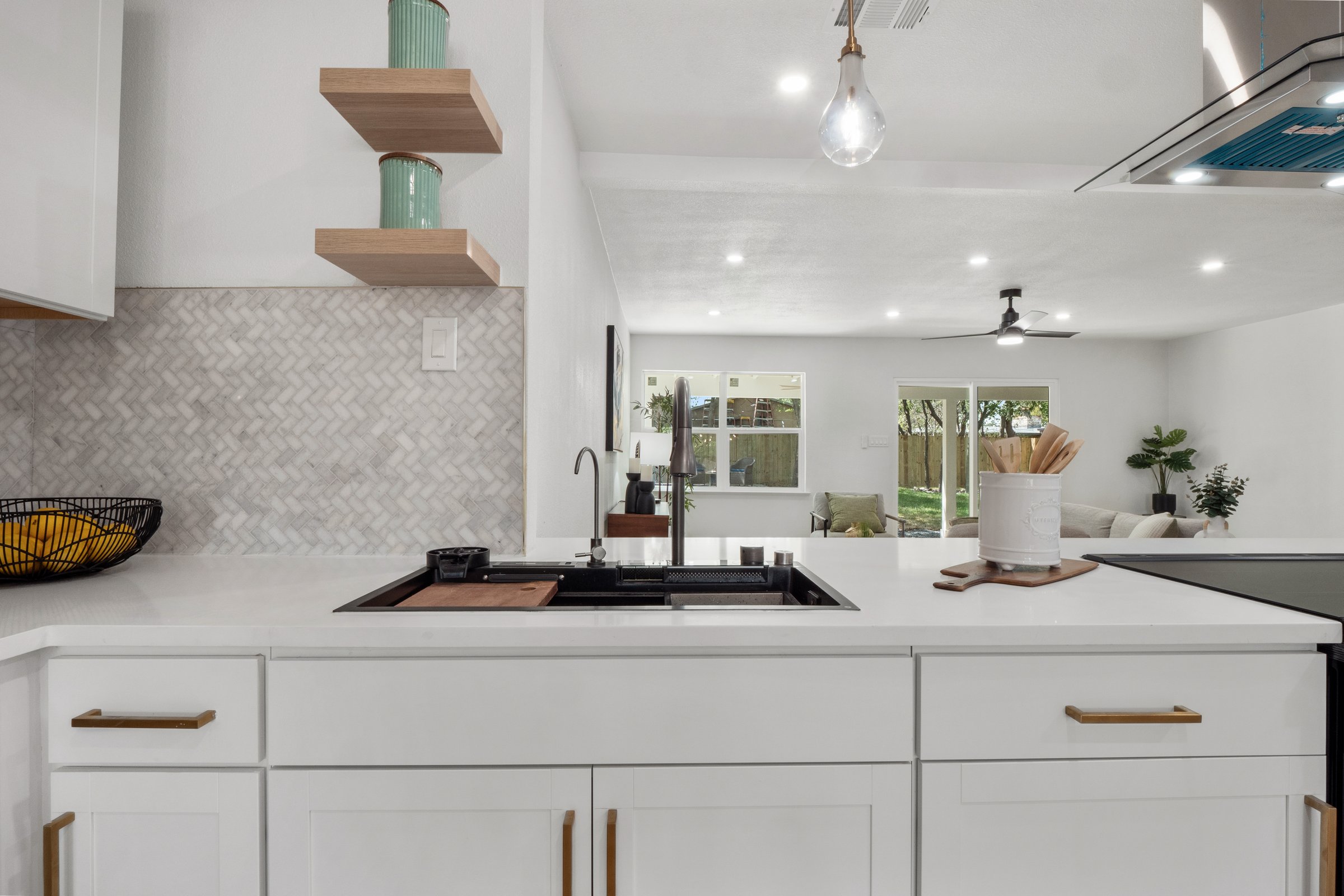 Modern kitchen with white cabinets, black sink, wooden shelves, and open living room with large windows in background.