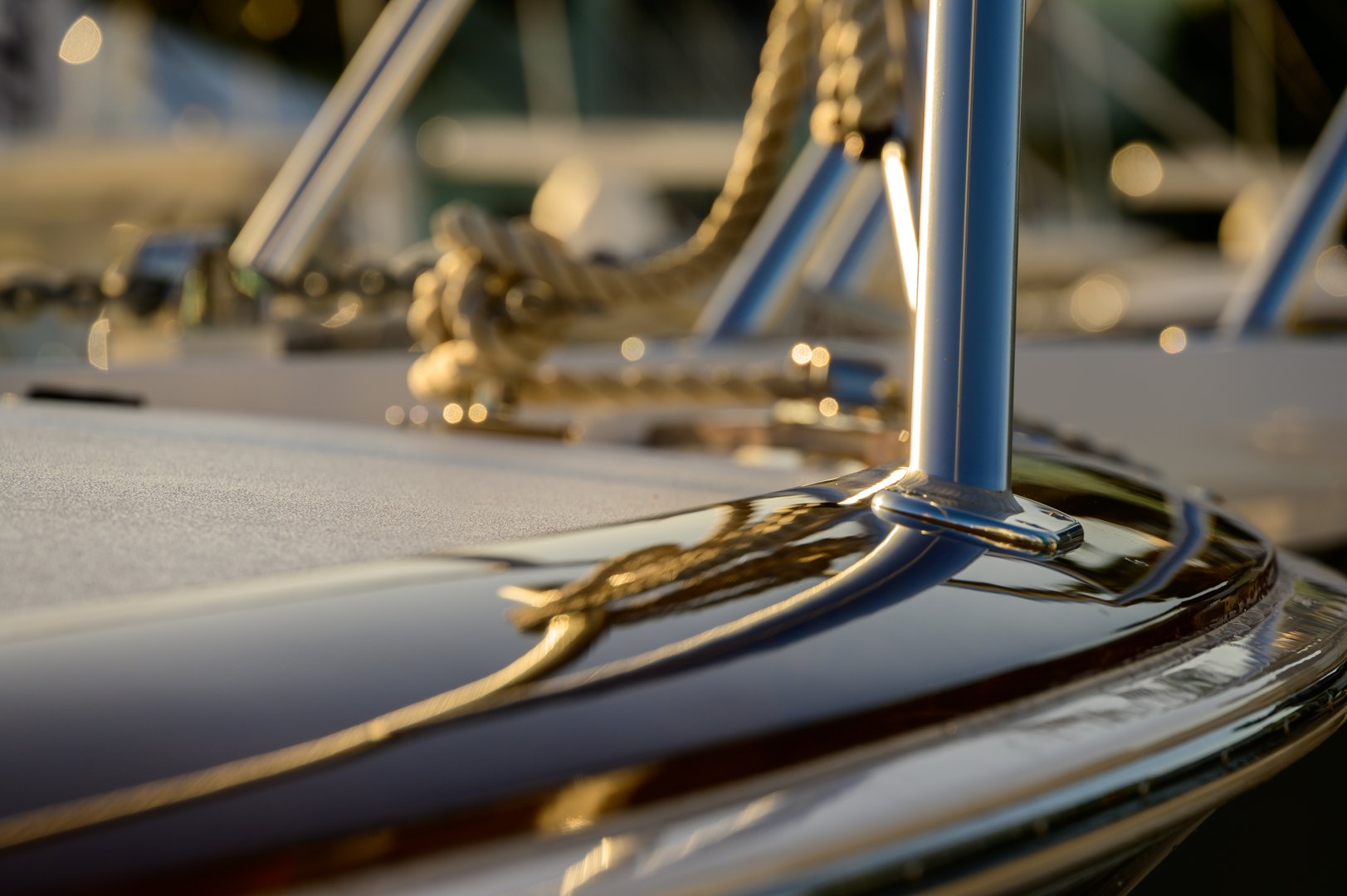 A close-up view of a glossy boat hull with a blue mast rising from the deck, ropes and hardware in soft focus. Sunlight creates golden reflections along the curved surface, conveying luxury, craftsmanship, and maritime glamour at a prestigious boat show.