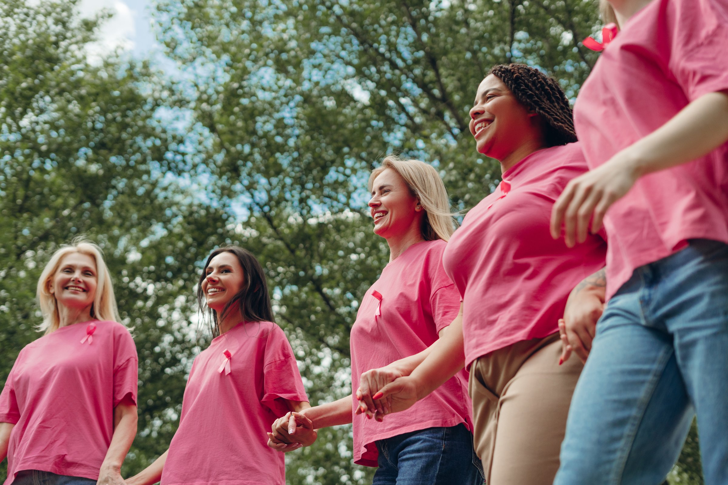 Multi-ethnic women of different ages, wearing pink t-shirts with pink ribbons pinned, hold hands and walk together outdoors, participating in breast cancer awareness event