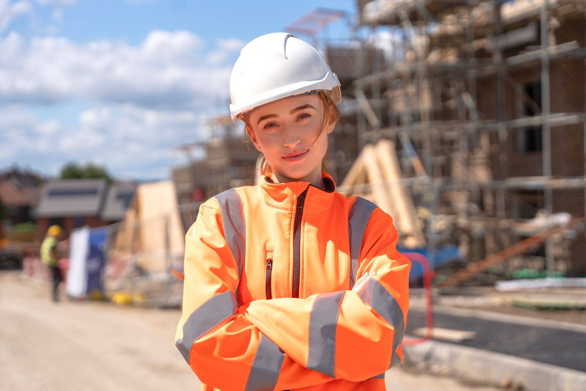 A young woman wearing a hard hat and reflective jacket stands with crossed arms at a construction site, showcasing confidence amidst ongoing building work on a clear day.
