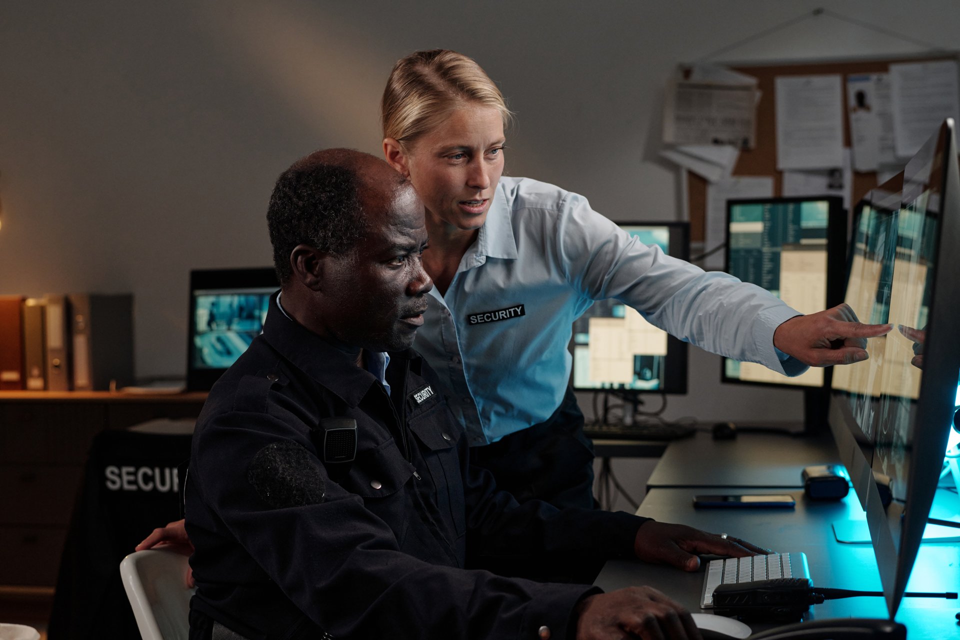 Young confident female security guard standing next to male colleague and pointing at computer screen while both watching cctv