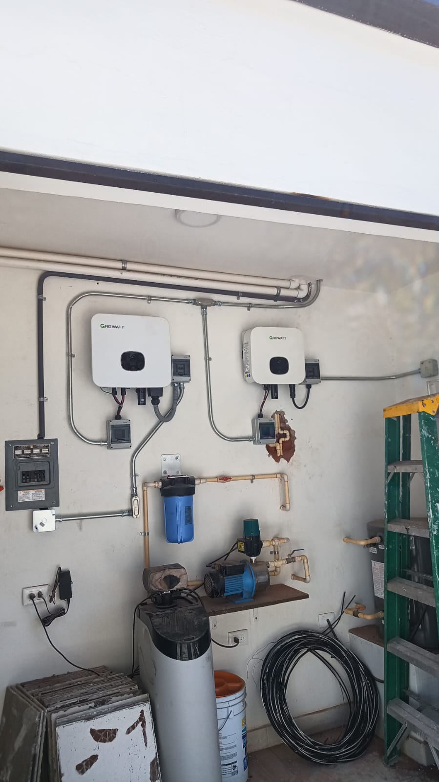 Electrical equipment and pipes mounted on a wall, with two inverters, a water pump, and a ladder nearby.