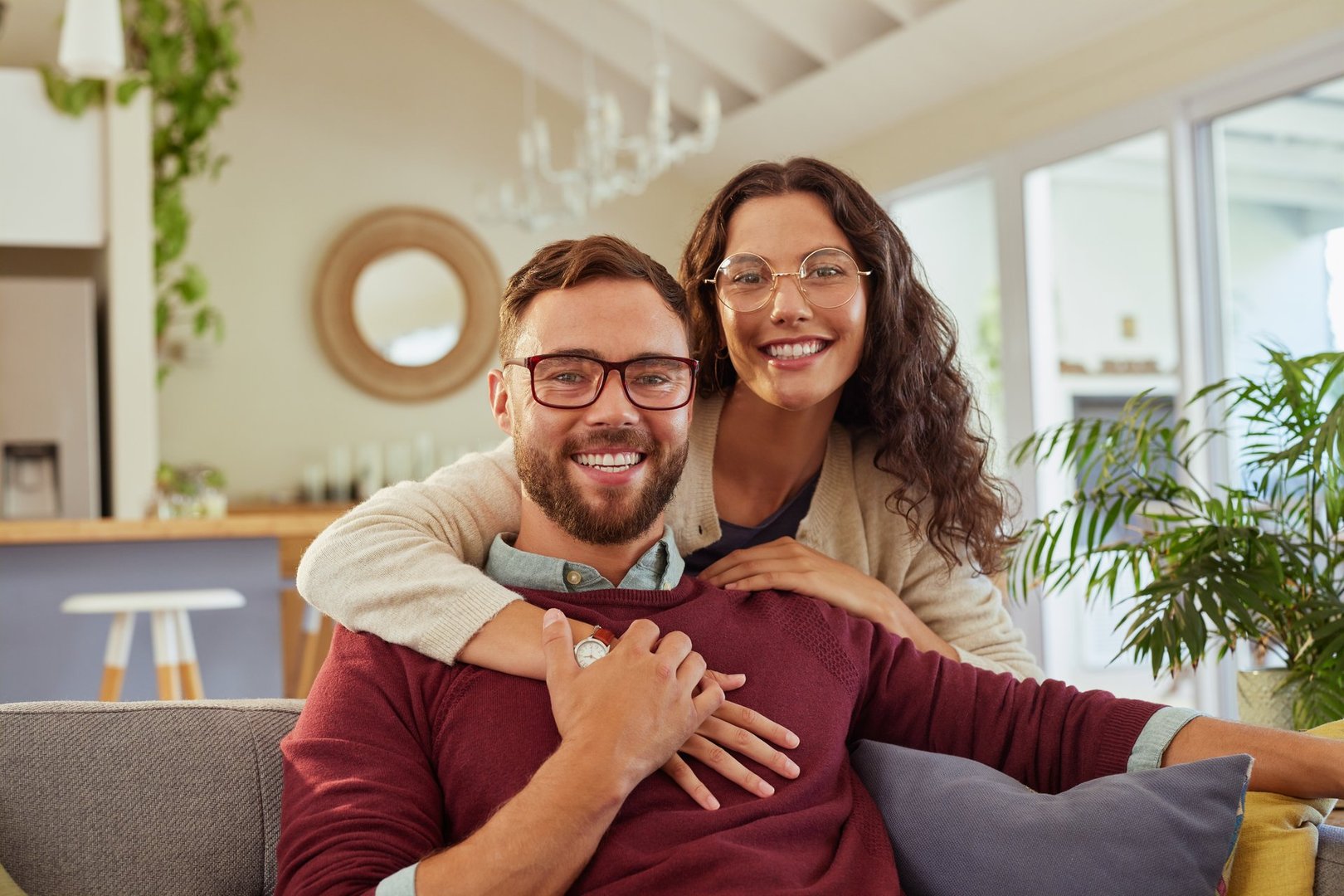 Happy smiling woman hugging husband with eyeglass from back while sitting on sofa at home. Mid adult couple in  love in cozy living room with a big grin. Portrait of satisfied man and beautiful wife embracing at home.