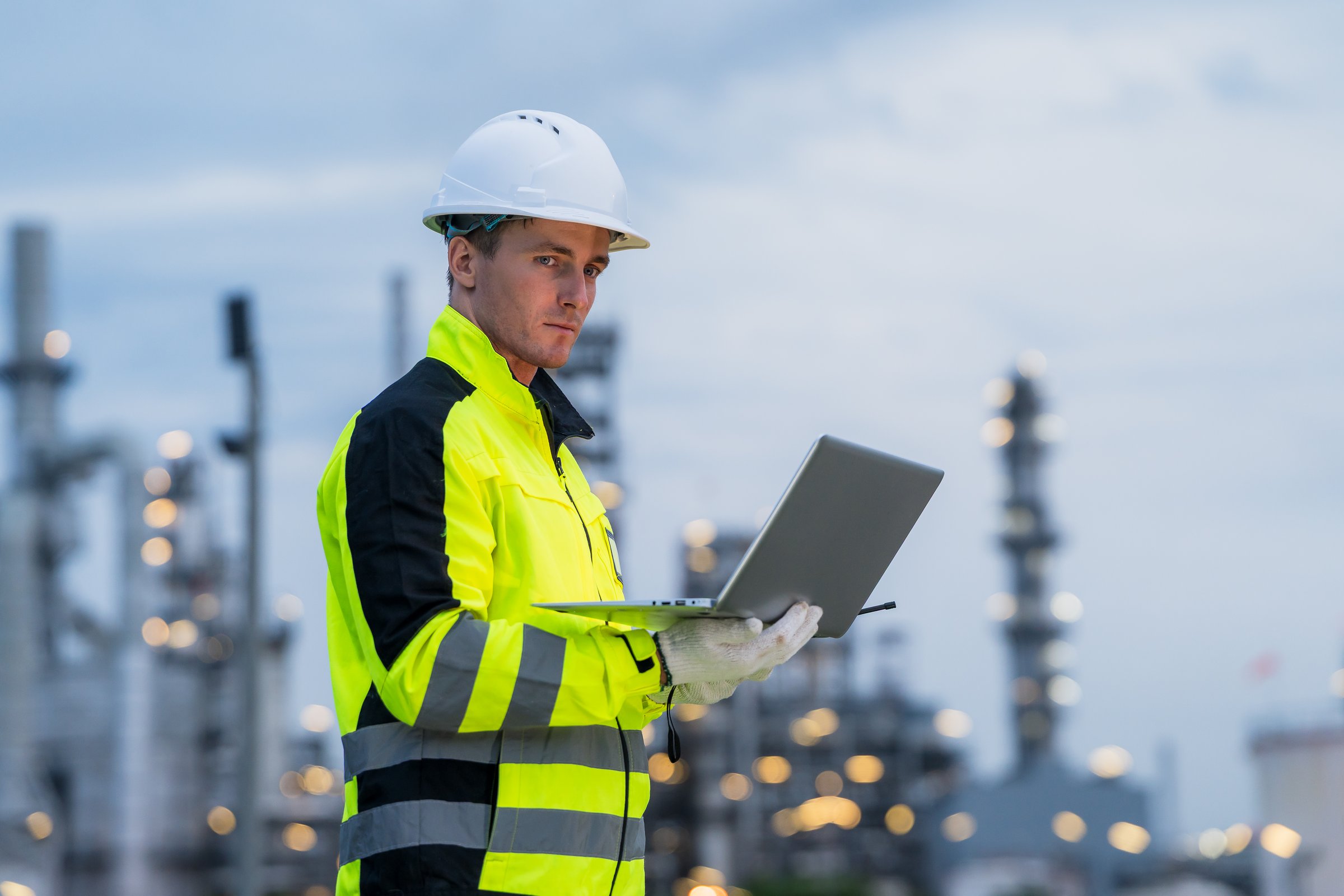 Engineer working on a laptop at a petrochemical industrial plant, monitoring the system during the night shift.