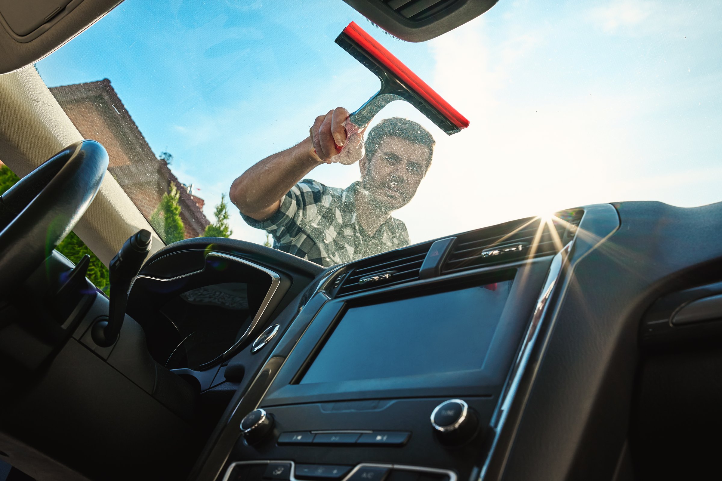 Male washing windshield in sunlight, view from from car inside. Man cleaning car windshield from outside using squeegee. Concept of car care, cleaning services and automotive hygiene
