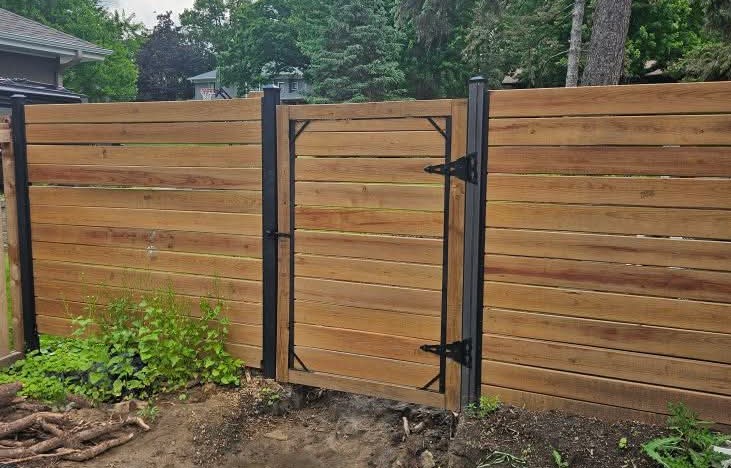 Wooden fence with black metal hinges and latch, surrounded by leafy plants and trees in a suburban setting.