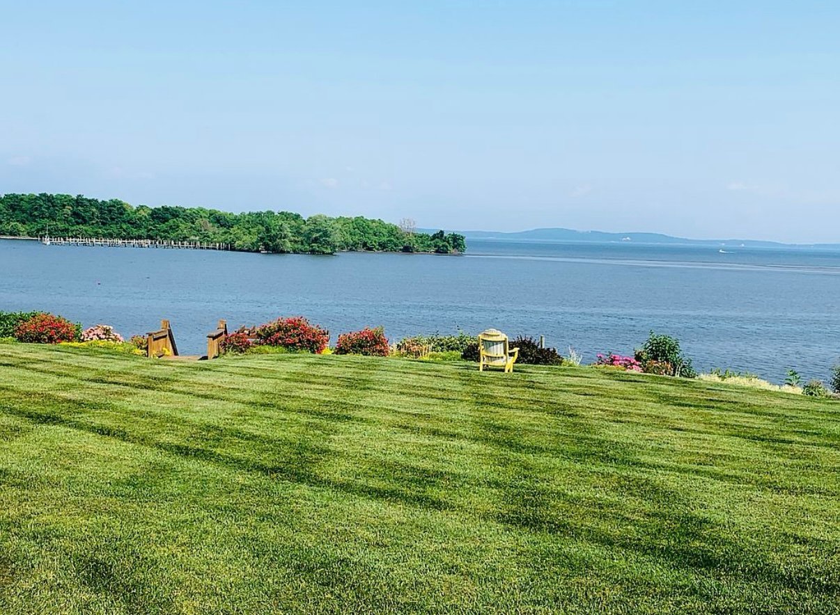 Lawn with Adirondack chairs overlooking a calm lake with distant tree-covered shoreline under a clear blue sky.