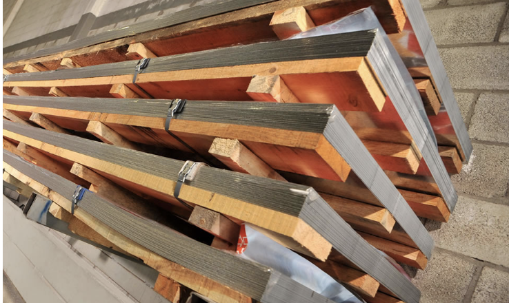 Stack of metal sheets bound with wooden spacers, leaning against a brick wall in a warehouse.