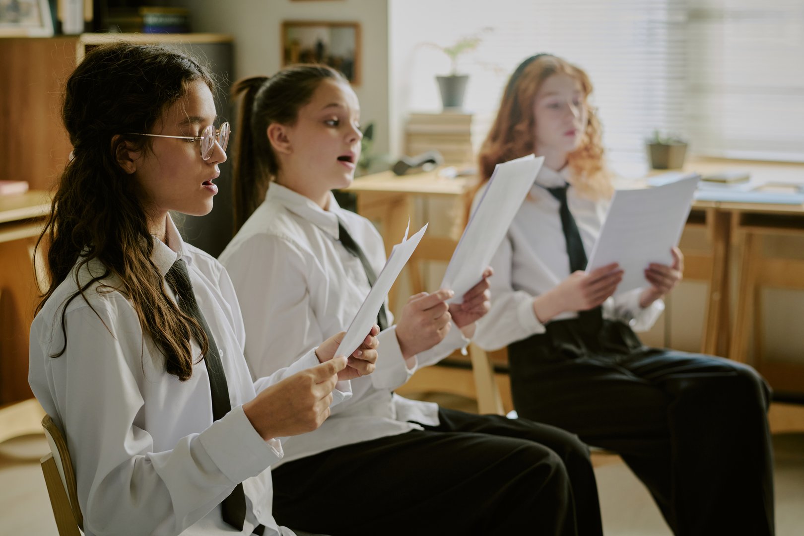 Group of students engaging in singing practice while holding sheets of paper in a music classroom setting with books and plants in background