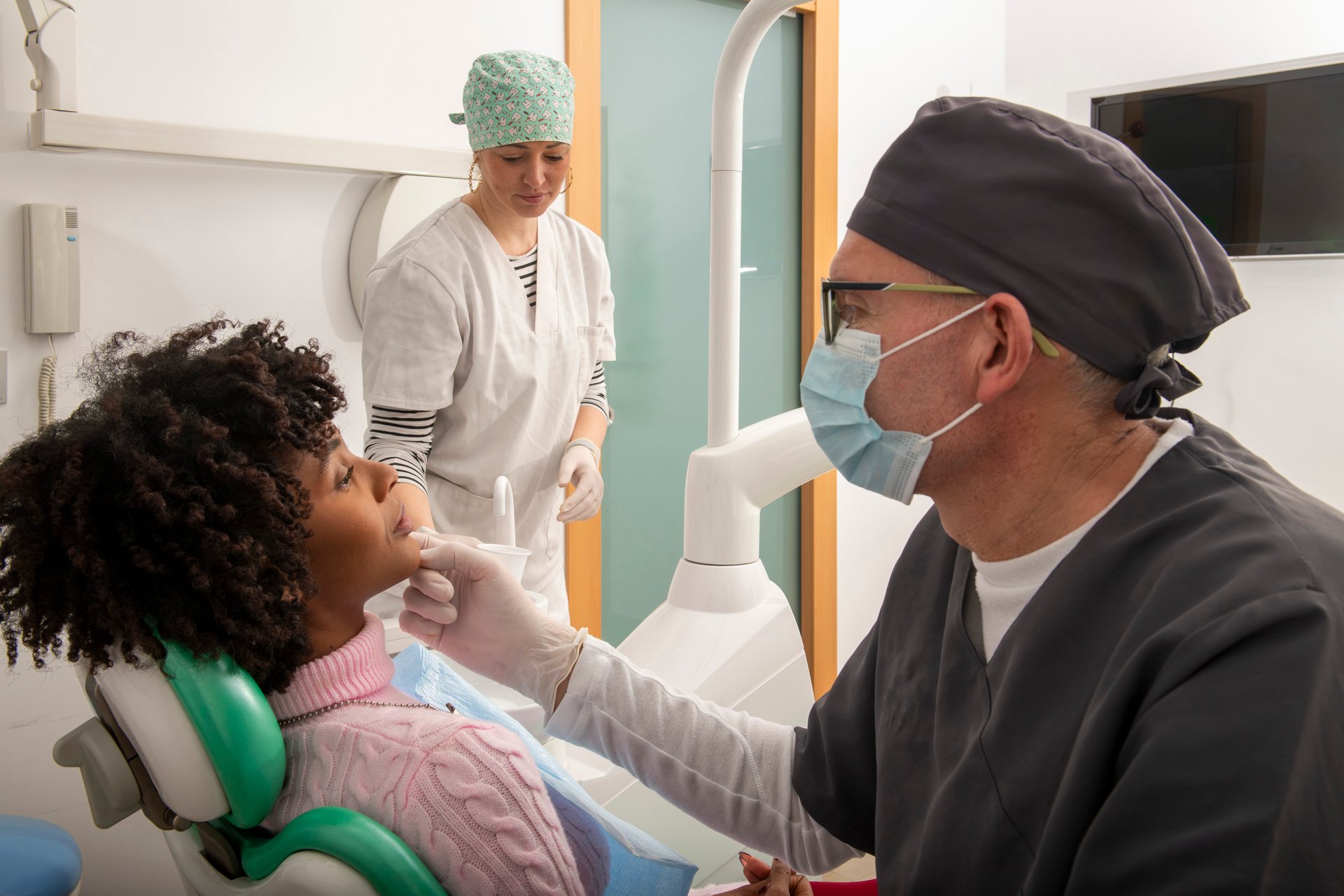 Dentist wearing mask and gloves examining patient's teeth while dental assistant prepares tools in a dental clinic