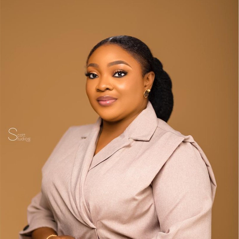 Woman in a light gray blazer posing against a beige background, with her hair pulled back and wearing subtle makeup.