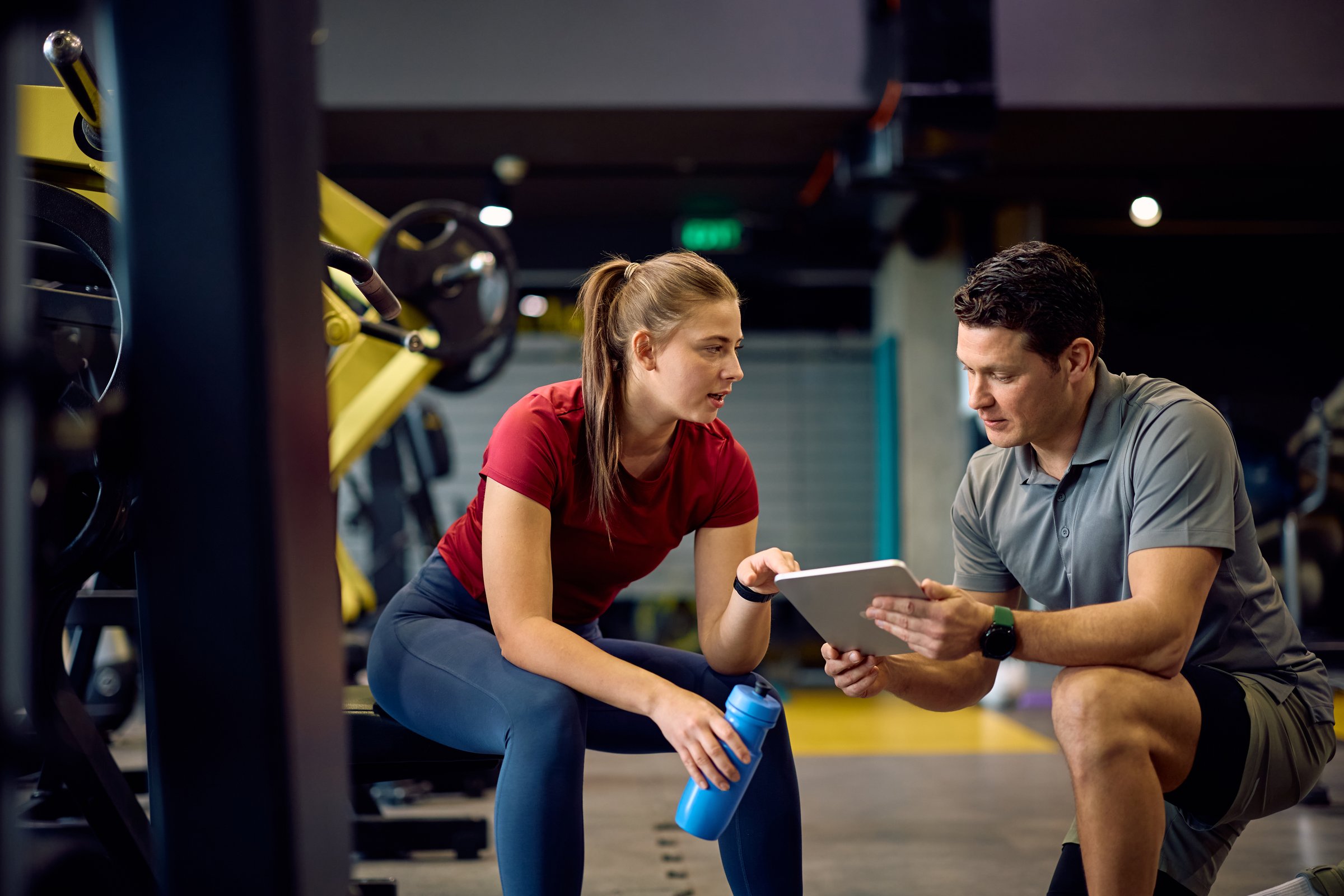 Young athlete and her fitness instructor analyzing plans on touchpad during sports training in a gym.