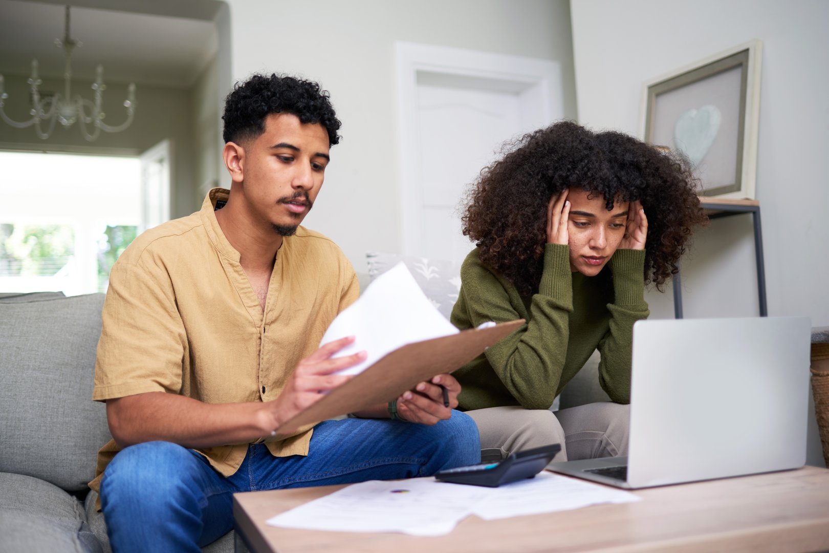 A couple sitting on the sofa in their living room looks concerned as they review financial paperwork and work on a laptop, reflecting challenges in budgeting and financial management.