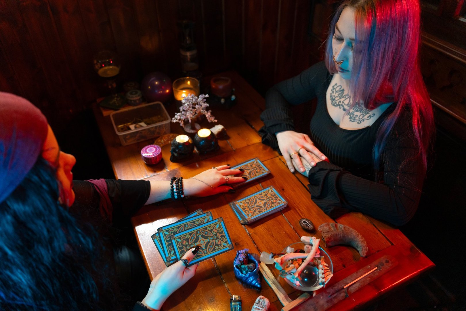 Fortune teller reading tarot cards spread on wooden table to young woman with colorful hair in a room with mystical atmosphere