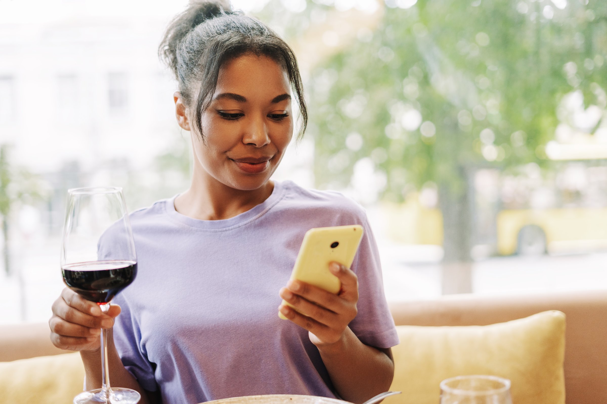 Happy African American woman enjoying wine and using smartphone in restaurant and holding glass with red wine, social media. Online technology concept