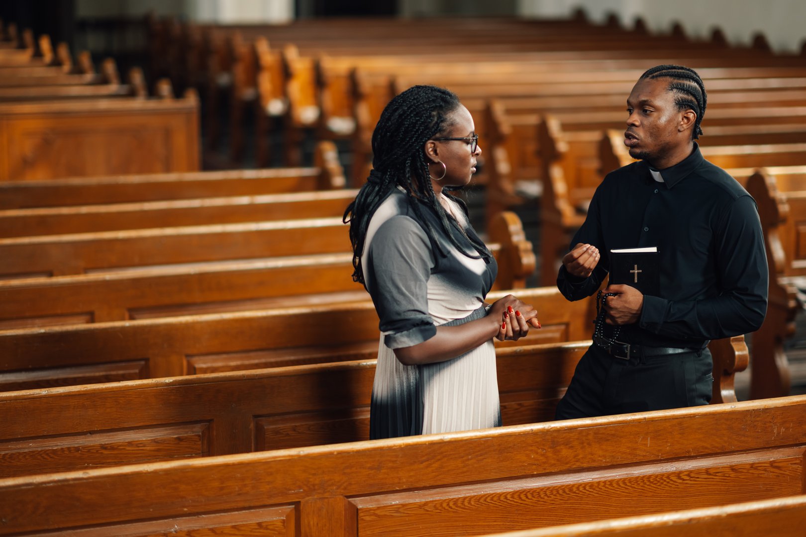 Priest is holding a bible while speaking with a woman in an empty church. They are standing in the church pews having a serious discussion