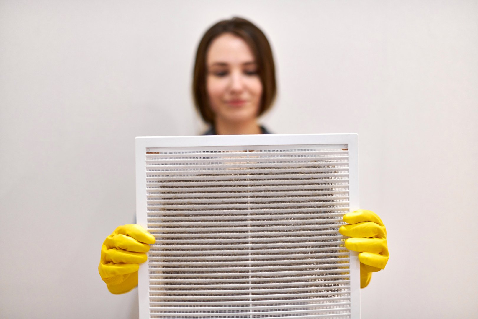 Woman holds ventilation grill with dust filter to clean it. Extremely dirty and dusty white plastic, harmful for health. Housewife in protective yellow rubber gloves and blue-grey uniform, blurred.