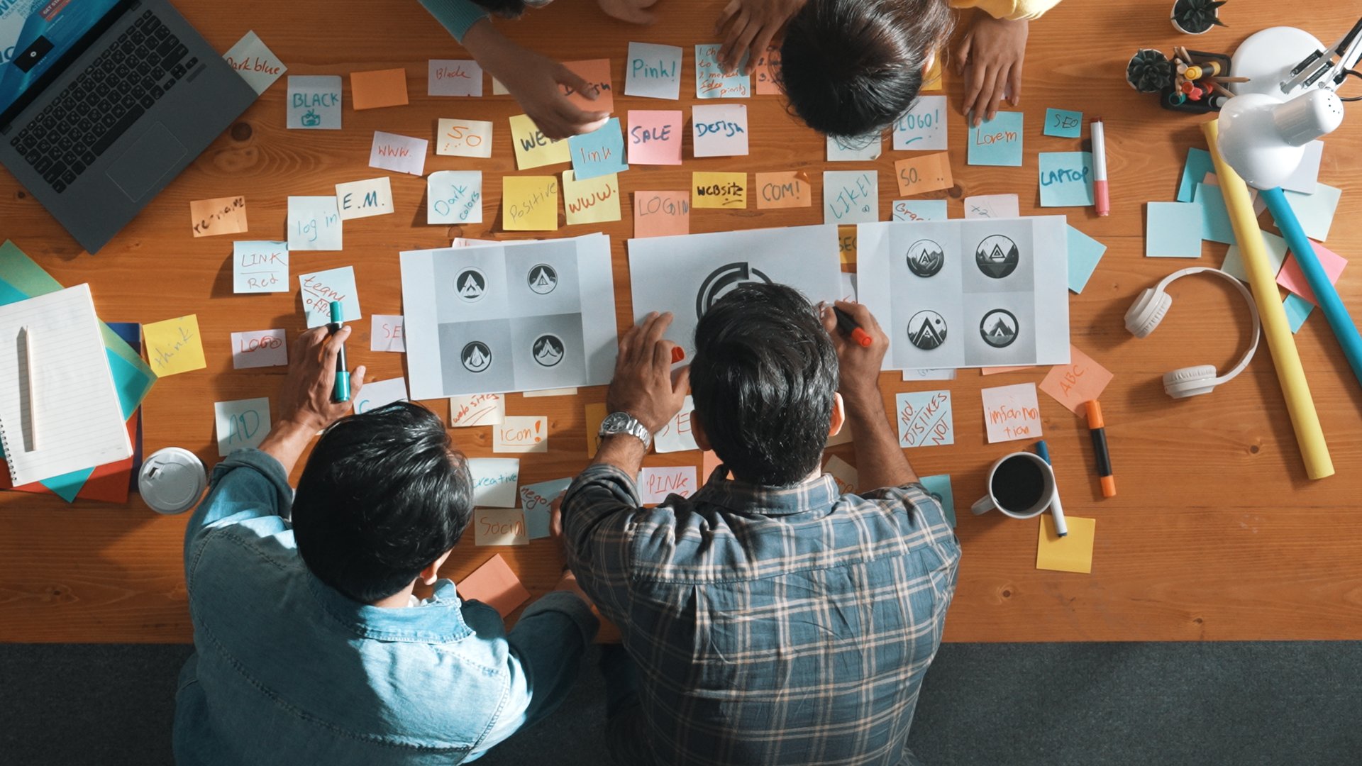 Top down aerial view of skilled designer hold logo while discuss with team. Aerial business group working together sharing idea about logo graphic design by using sticky notes and tablet. Symposium.