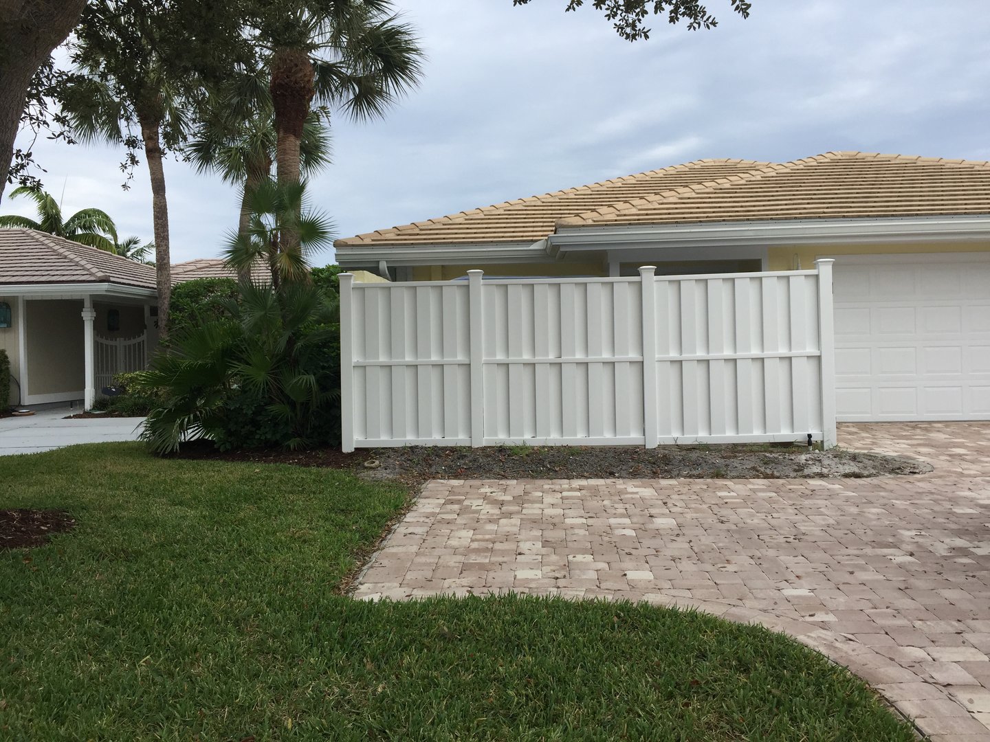 A suburban house with a white fenced driveway, beige roof, and landscaped yard featuring palm trees and grass.