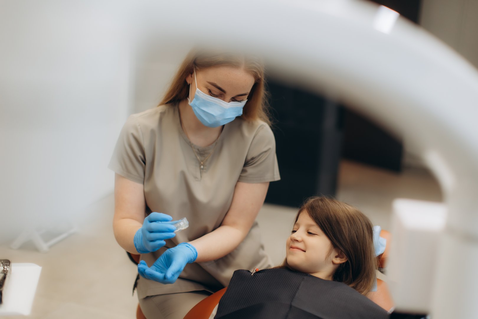 Female dentist wearing mask and gloves preparing filling material for little girl in dental chair. Dentist shows aligners, braces, caps