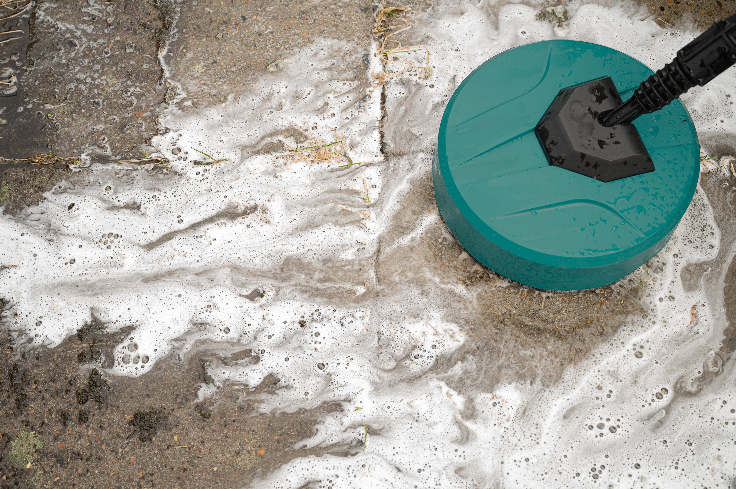 round high-pressure washer spraying foaming water on cement tiles overgrown with grass and dirt