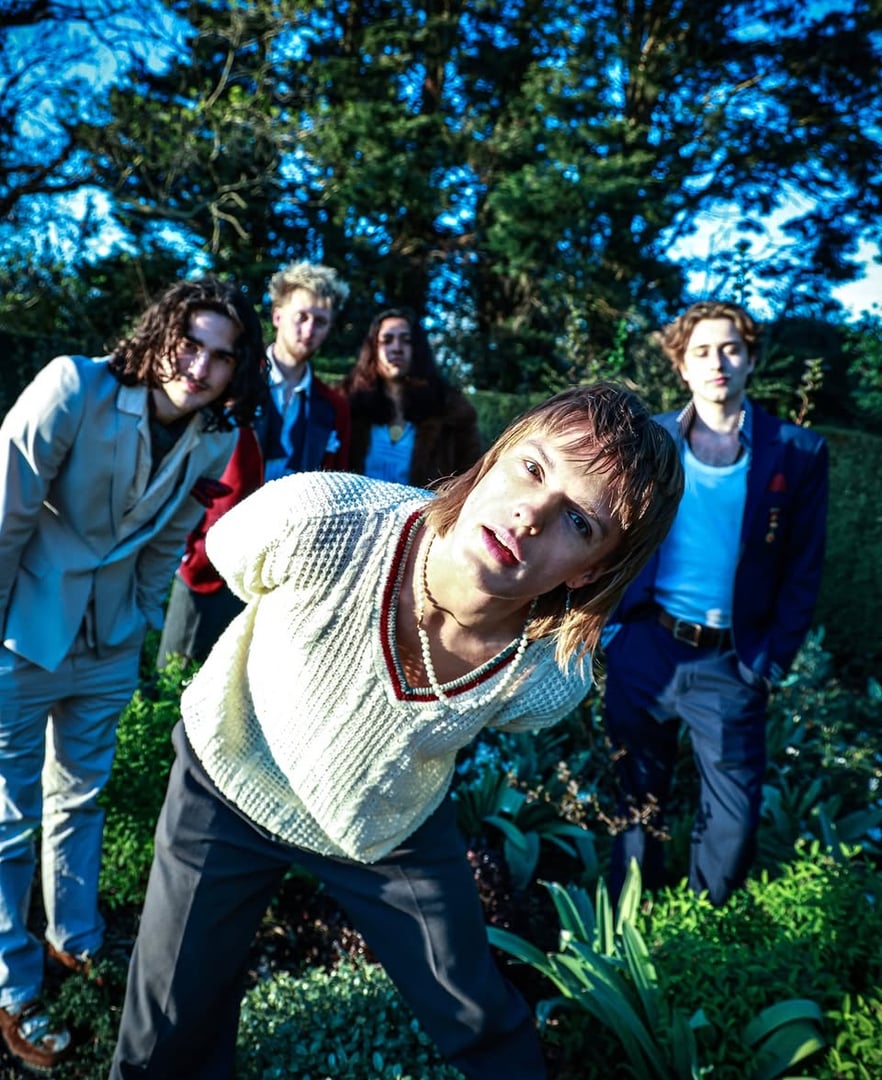 Group of five young adults posing outdoors, surrounded by greenery, with one person leaning forward toward the camera.