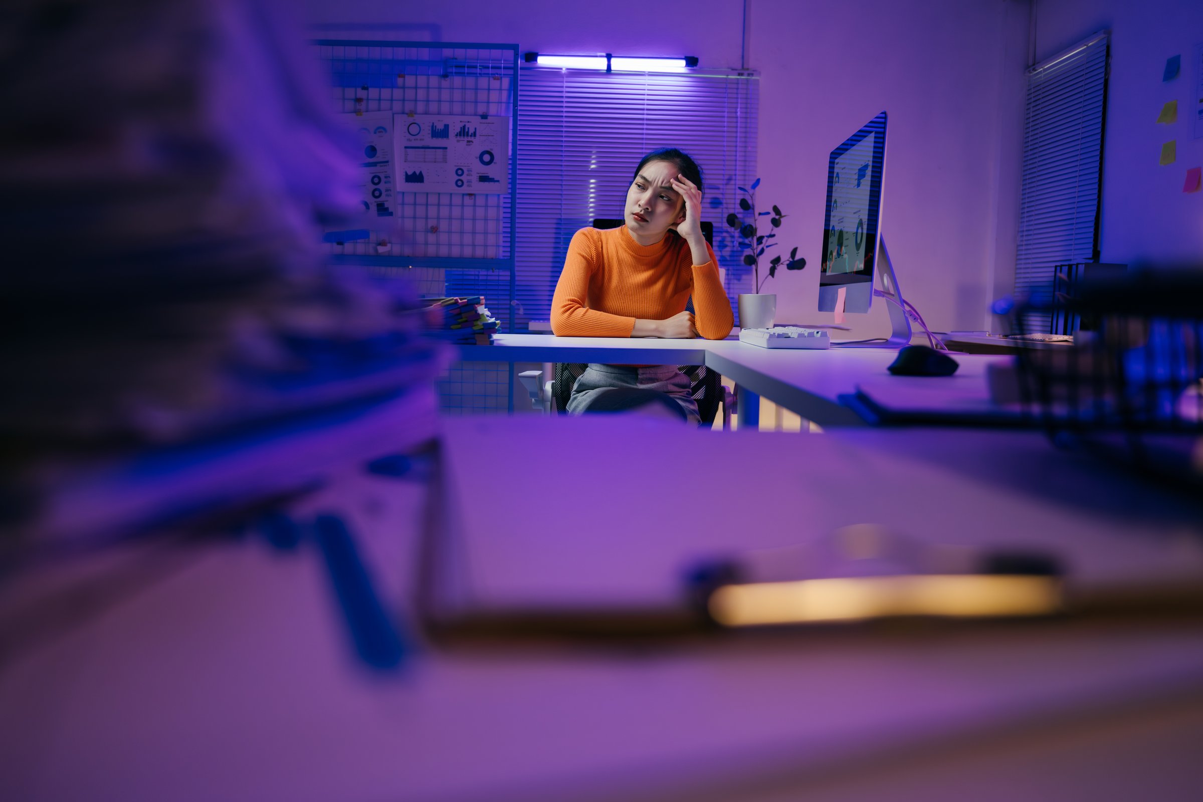 Tired business woman sitting at office desk with computer and paperwork, holding her head in her hand, suffering from a headache in a dark office with purple neon light