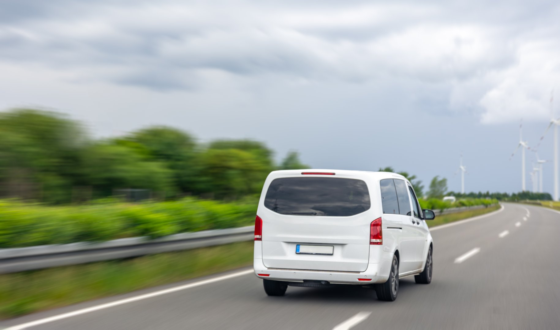 A minivan driving along a scenic highway with greenery and wind turbines in the background. Motion blur enhances the speed feel.