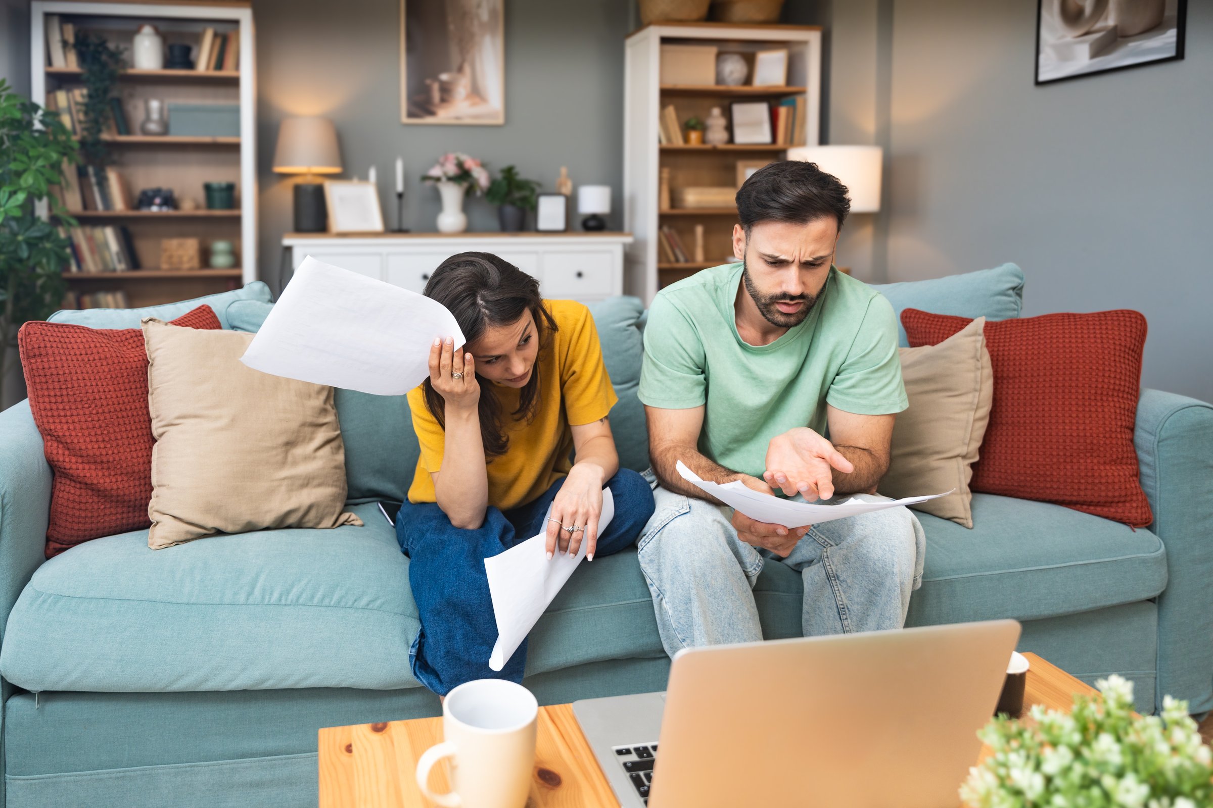 Worried young couple sitting on sofa at home, analyzing financial documents and statistics on laptop and paper, stressed about unexpected expenses, debt, or negative budget report results.